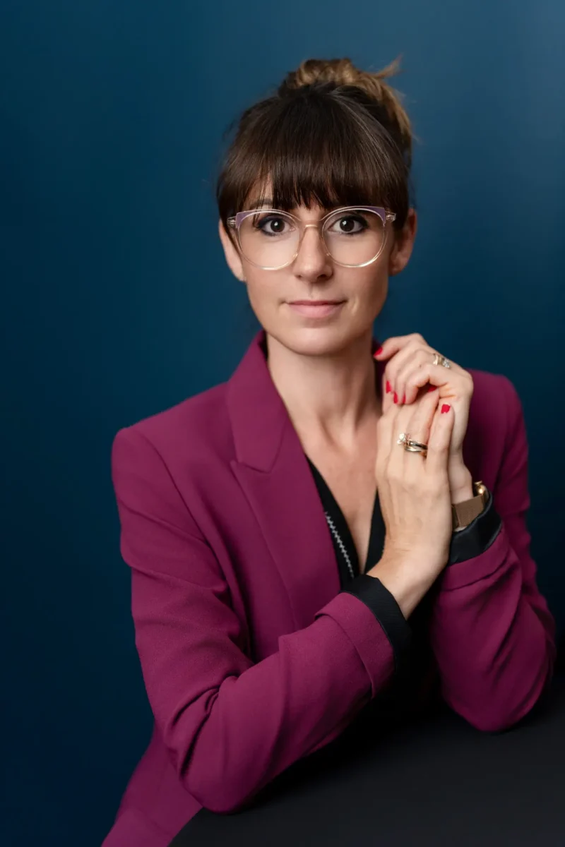 Portrait LinkedIn vs portrait, une femme pose devant un fond coloré bleu, porte une veste prune et des lunettes, image professionnelle adaptée au choix du portrait.