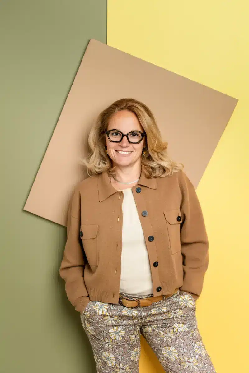 Portrait LinkedIn vs portrait, une femme souriante pose devant un fond coloré, porte une veste camel et des lunettes, image professionnelle adaptée au choix du portrait.